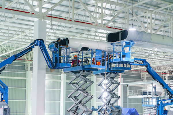Industrial contractors on scissor lifts safely installing large overhead ventilation ductwork to ensure optimal facility airflow.